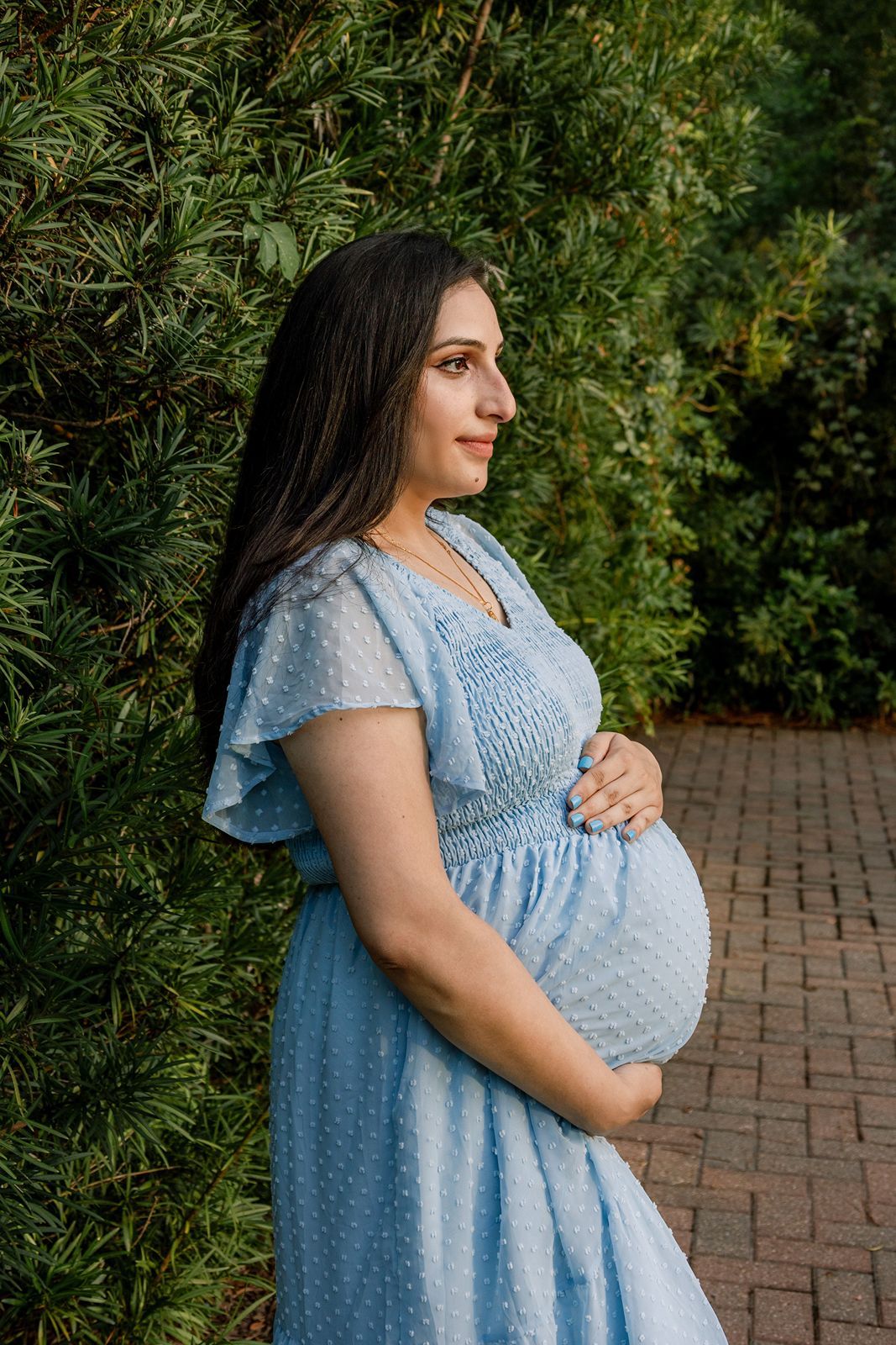 A pregnant woman in a light blue dress stands in a garden setting with brick pavers and lush greenery.