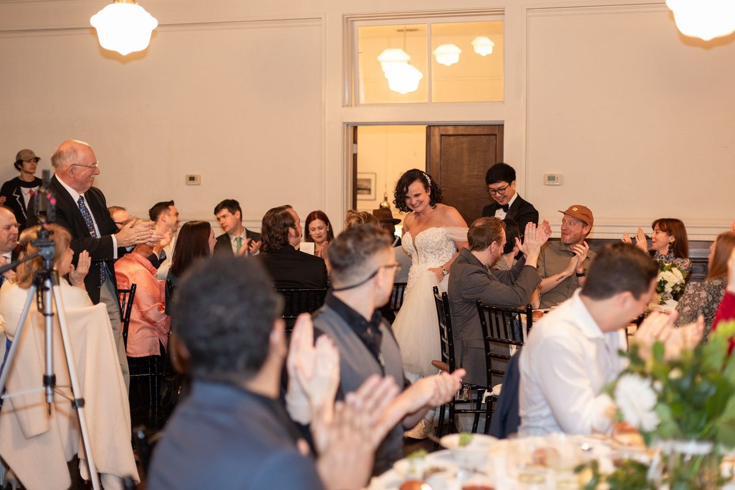 Wedding guests applaud and celebrate during an indoor reception ceremony with warm lighting.