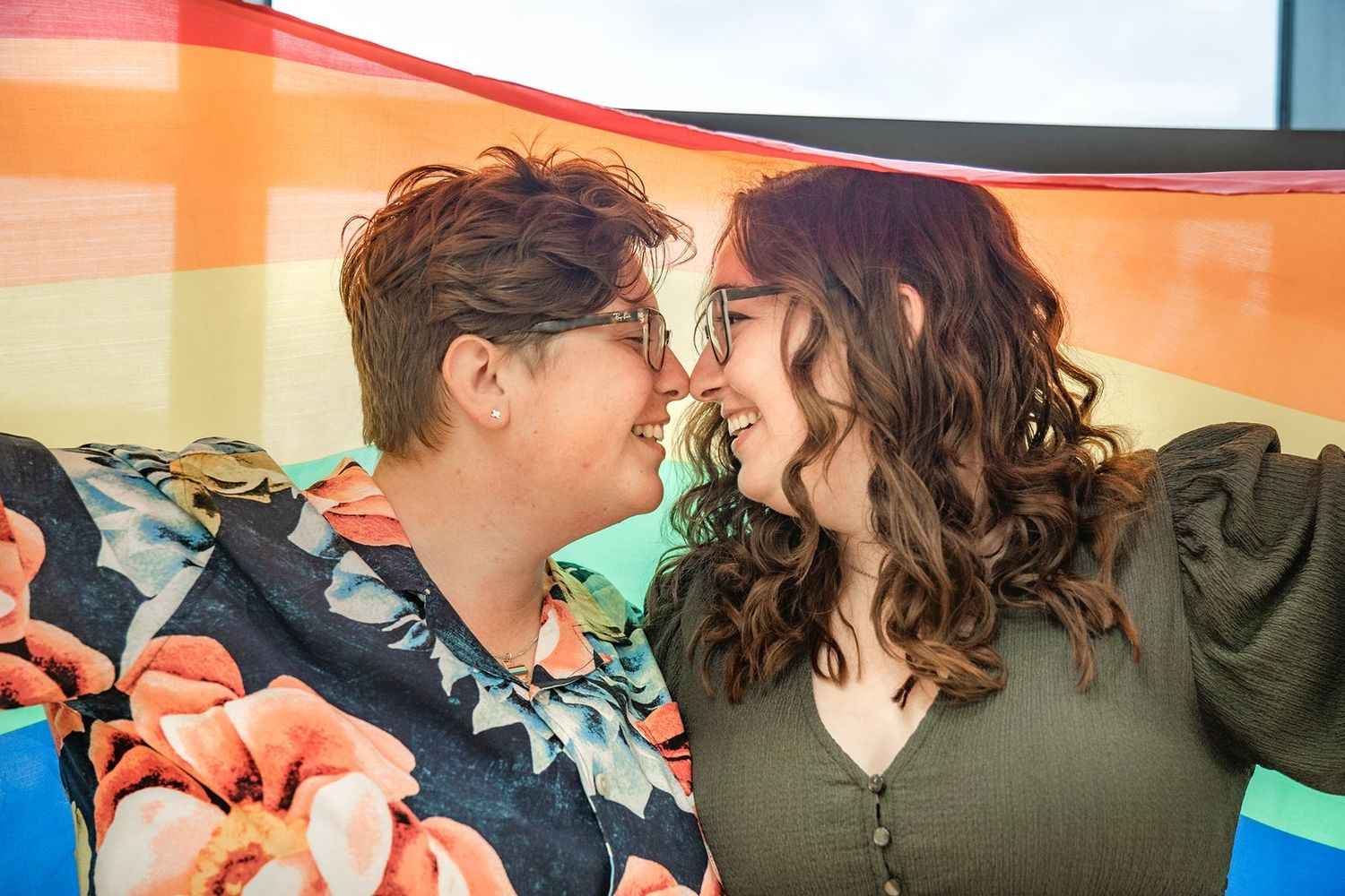 Two people share a tender moment while sitting together beneath a colorful rainbow backdrop.
