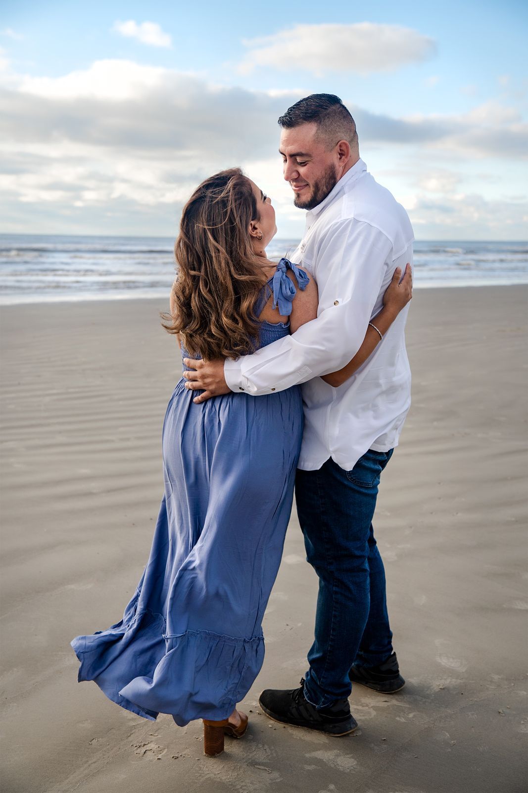 A romantic couple in blue and white outfits shares tender moments together on a cloudy beach at sunset.
