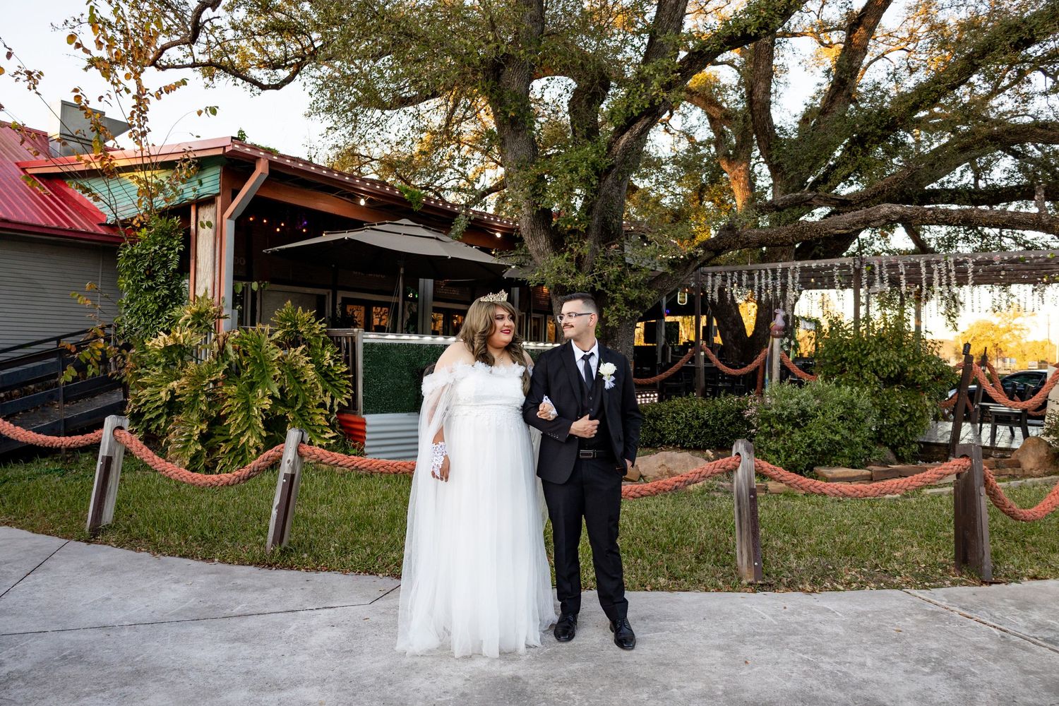 Bride and groom posing outdoors near a large oak tree at a rustic wedding venue with string lights.