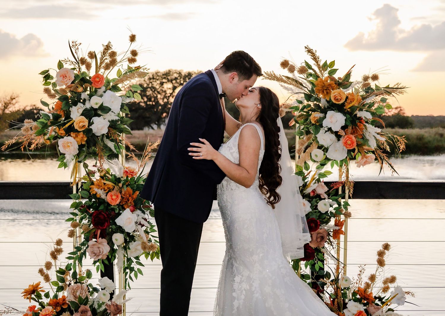 Bride and groom share first kiss under floral arch with orange and white blooms at sunset waterfront wedding ceremony.