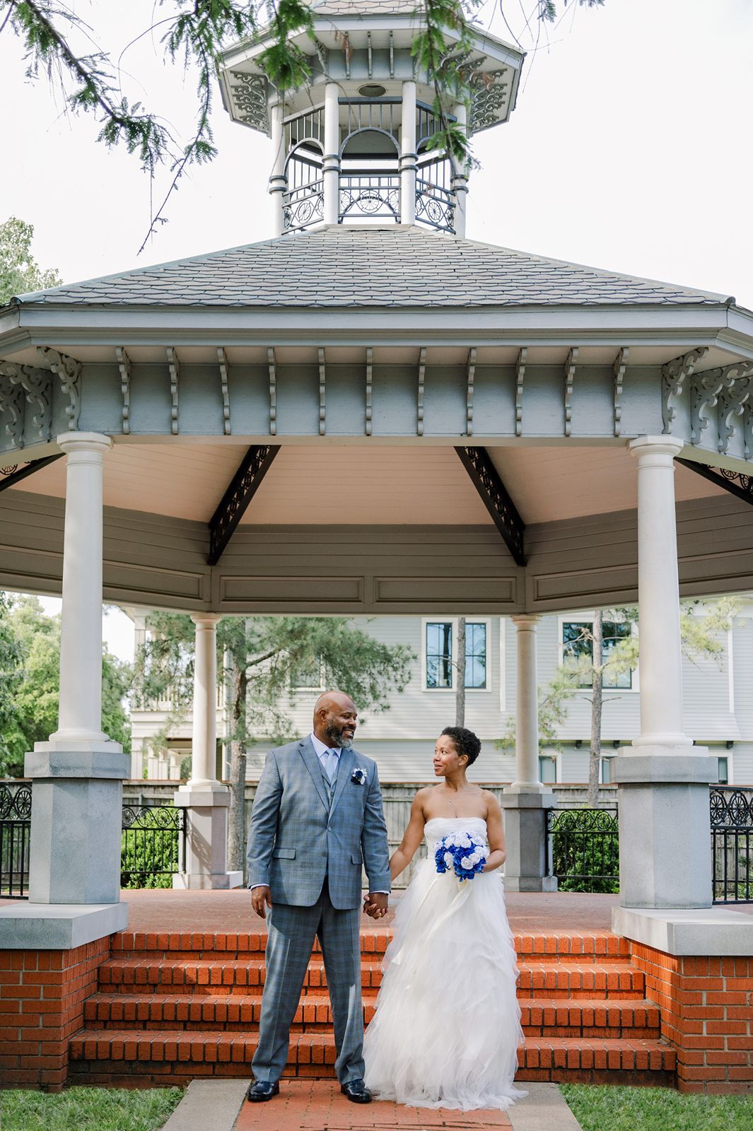 Bride and groom holding hands on brick steps in front of elegant white gazebo, bride holding blue bouquet.