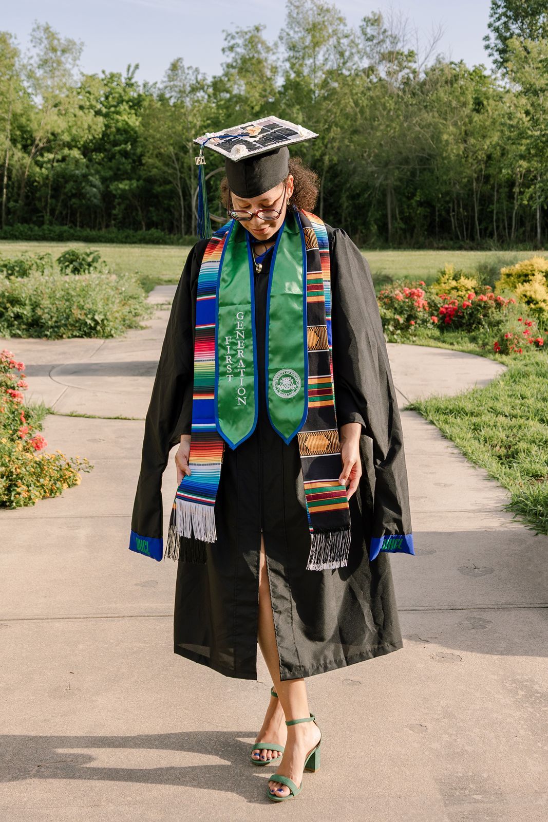 Graduate in black cap and gown with colorful stoles walks outdoors on a sunny day in a lush green park.