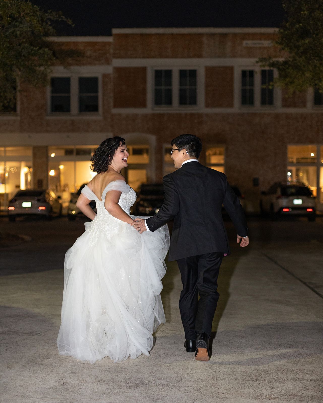 A couple in wedding attire runs joyfully across a courtyard at night with a brick building in the background.