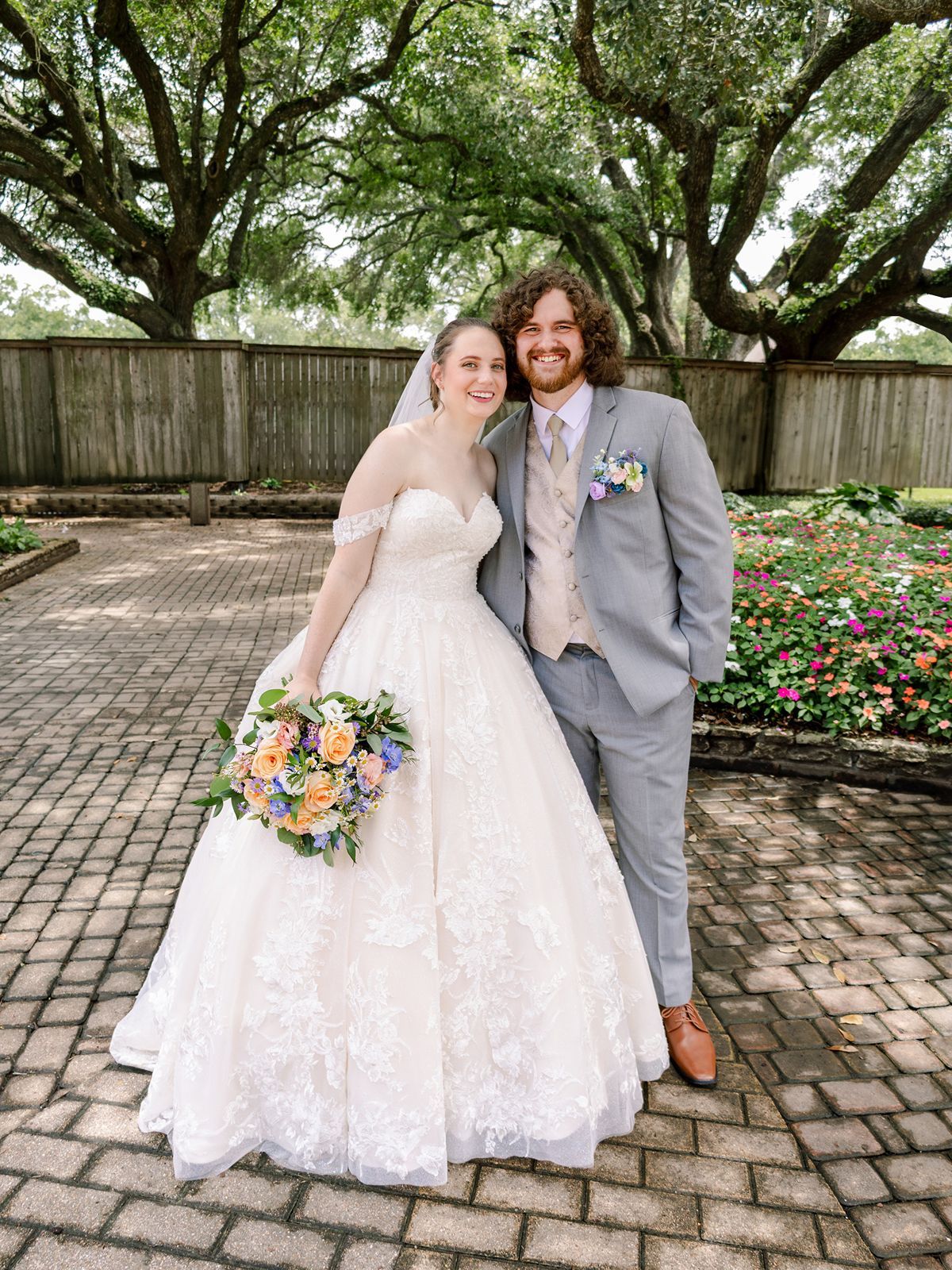 Couple in wedding attire sitting together on brick steps in a garden setting with colorful floral bouquet.