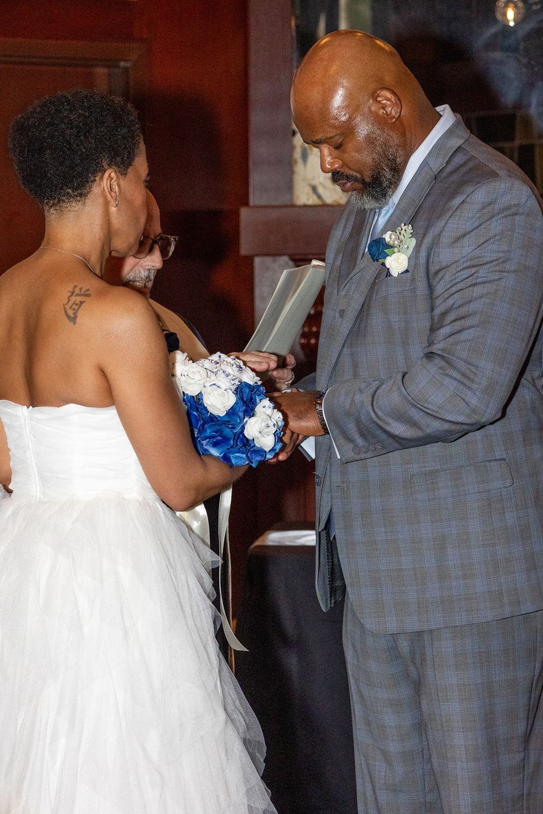 Bride and groom exchange rings during wedding ceremony, bride holds blue and white bouquet.