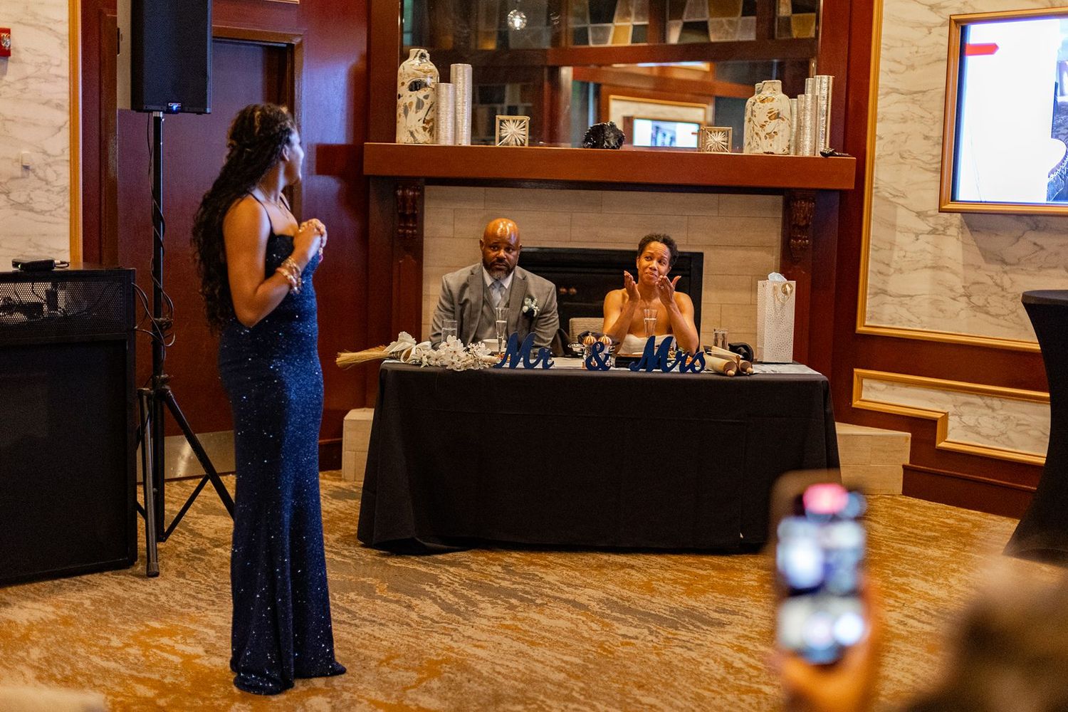 Woman in blue sequin gown speaking to couple seated at head table during reception.