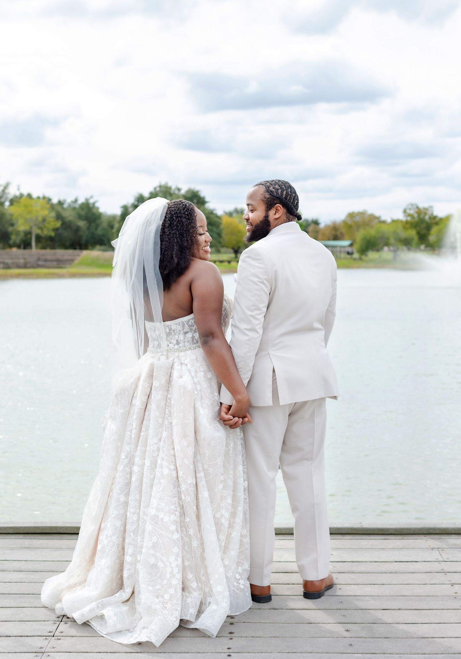 Wedding couple in white attire pose intimately on wooden dock by scenic lake.