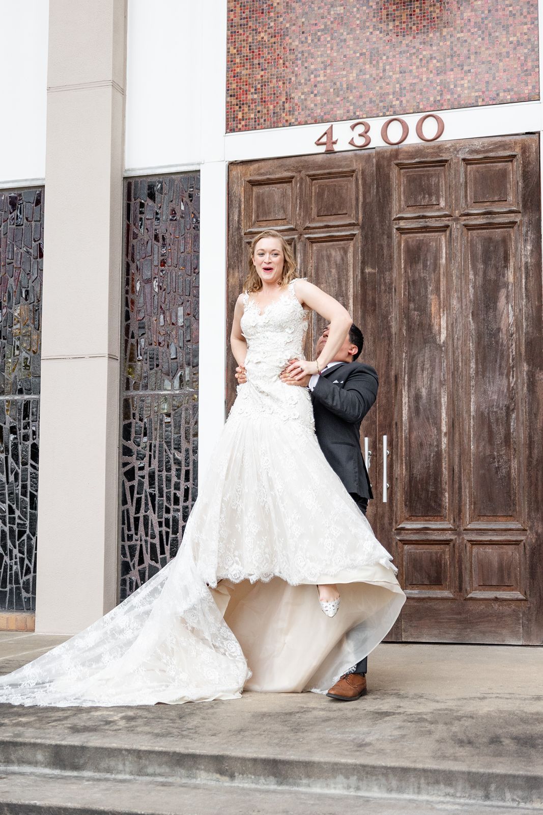 Playful groom lifting bride's wedding gown train outside wooden doors at 4300 venue.