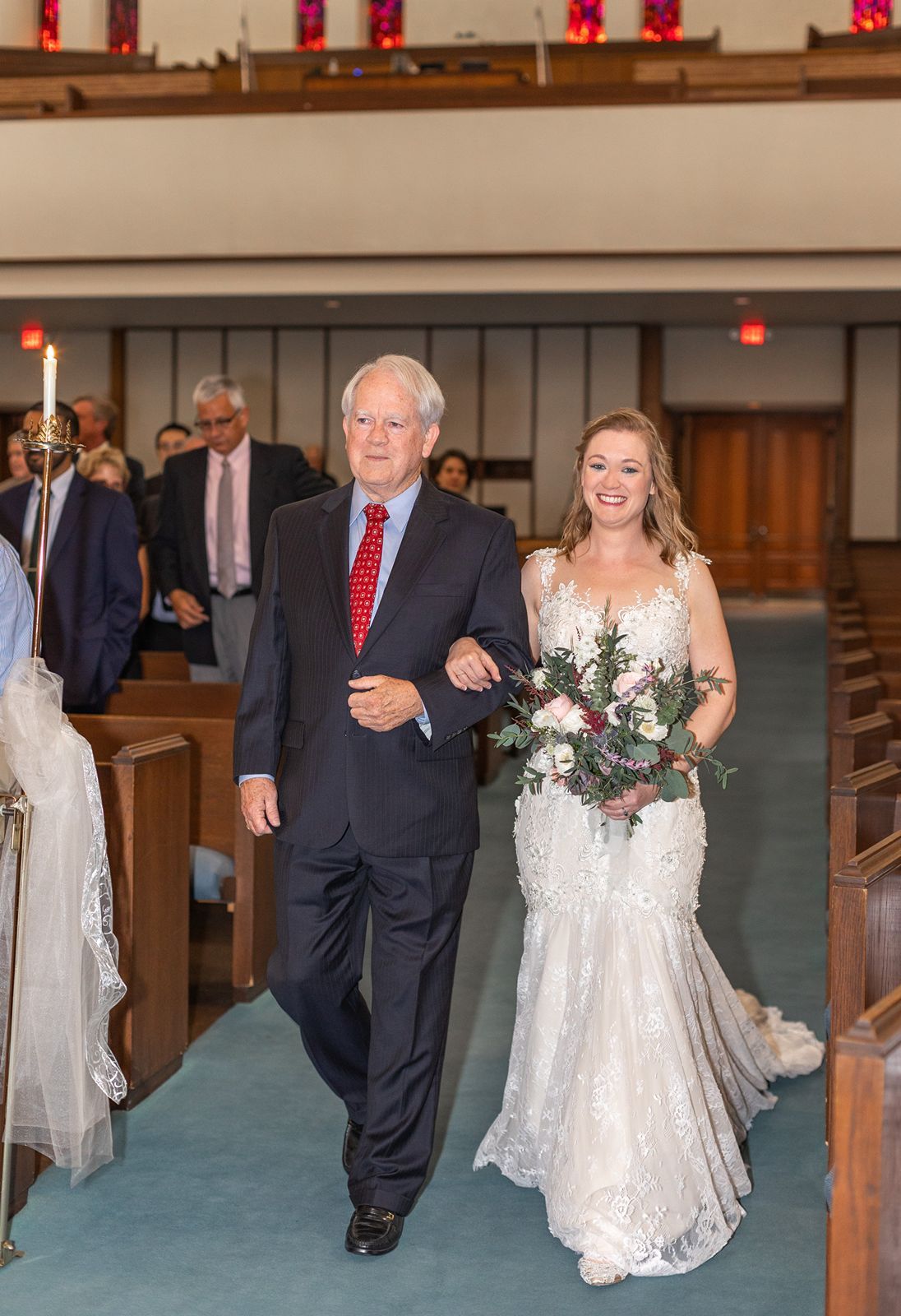 Bride in lace gown walks down church aisle with older gentleman in navy suit holding bouquet of pink flowers.