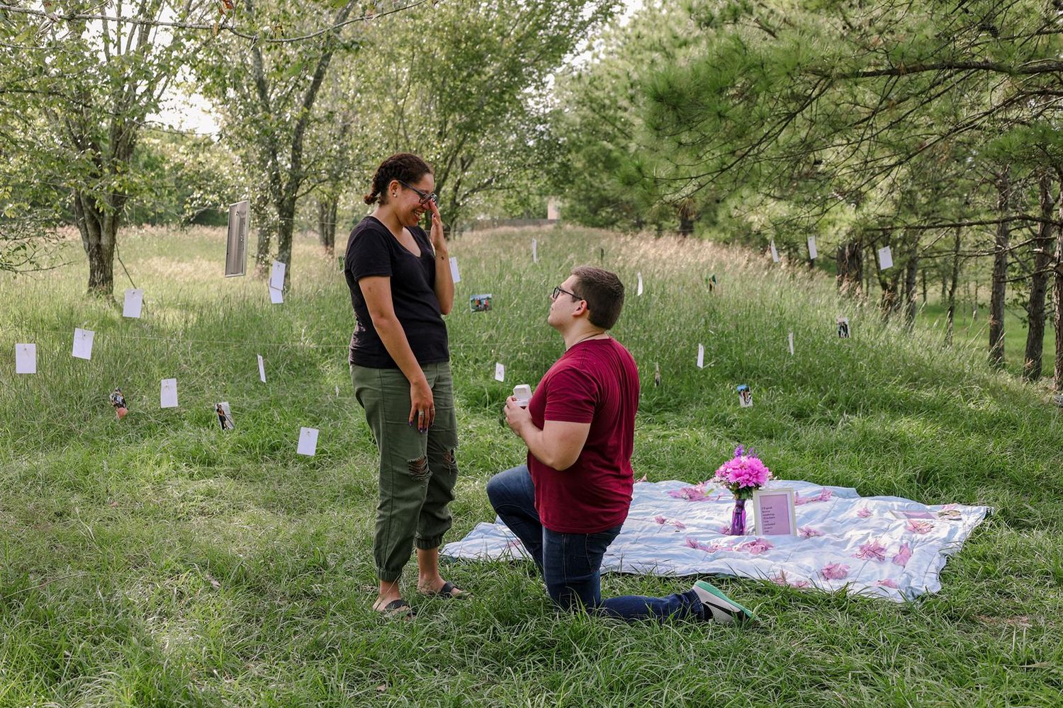 Person proposing on one knee to partner on floral blanket in lush green park setting with trees.