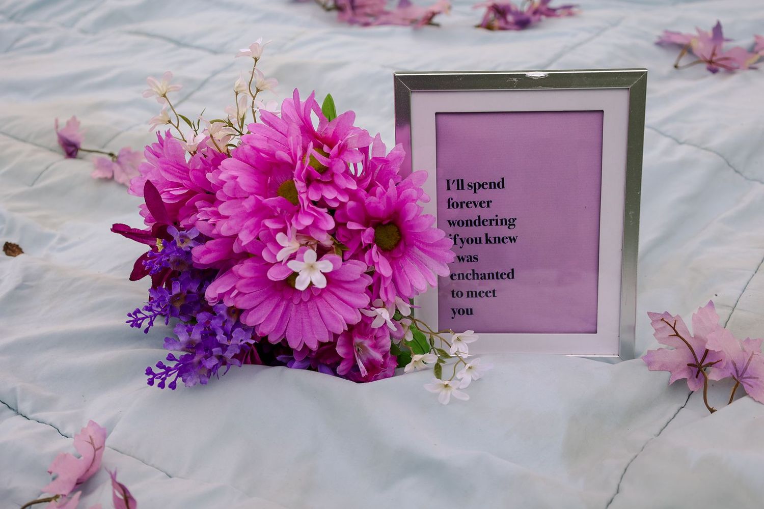 A vibrant pink flower bouquet beside a framed romantic quote on a white blanket with scattered petals.