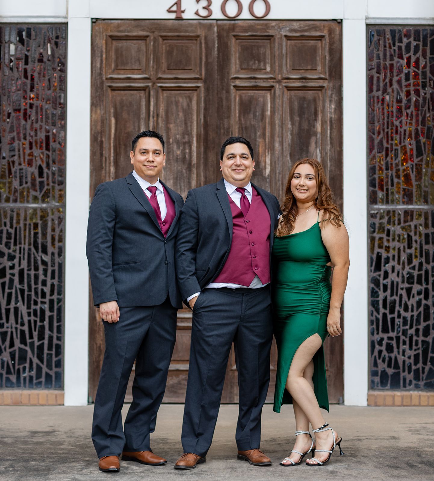Three formally dressed guests pose together outside building 4300, two men in suits and a woman in a green dress.