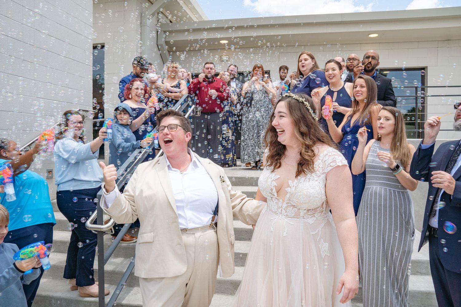 Joyful newlyweds exit wedding venue as guests celebrate with bubbles on sunny steps.