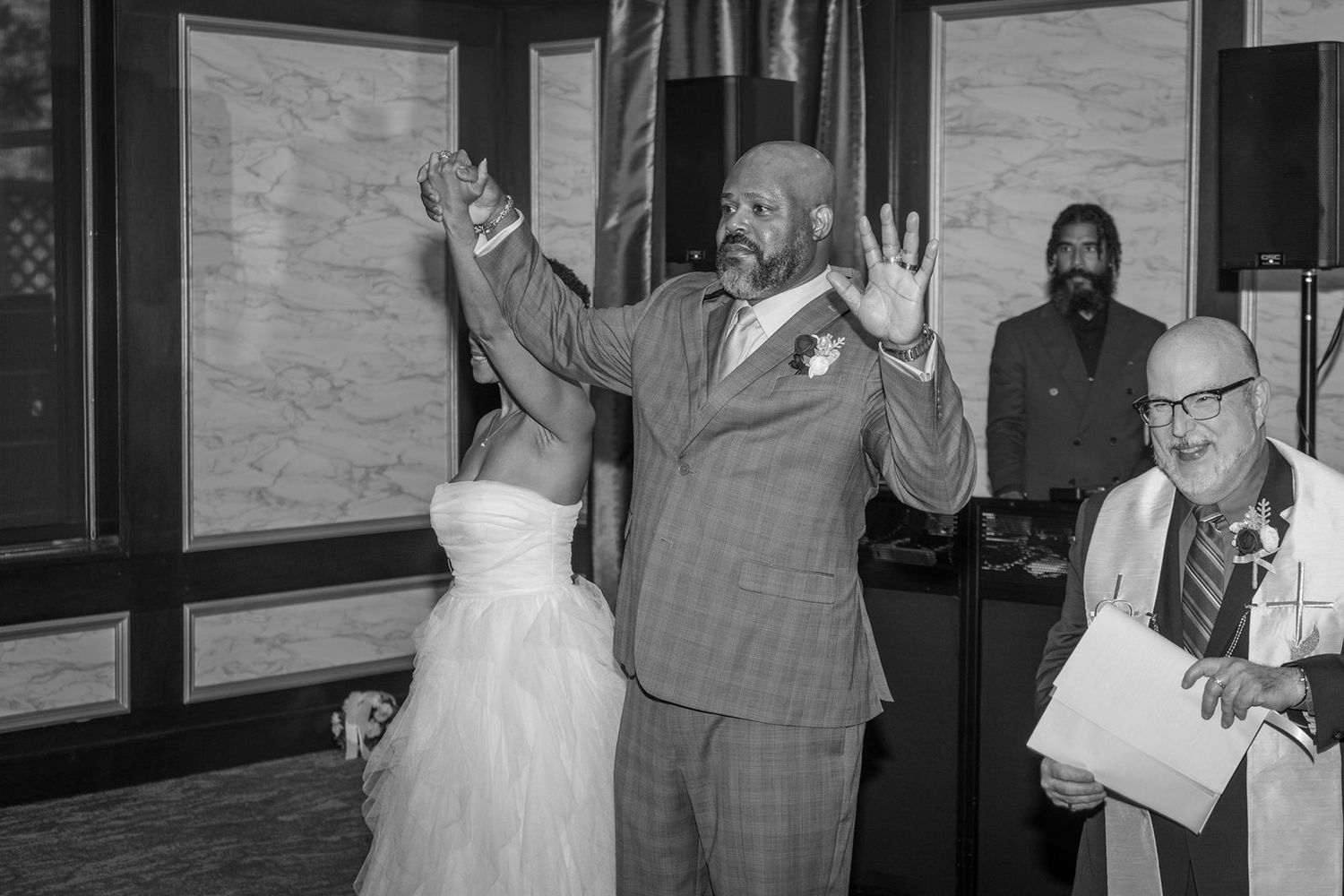 Joyful groom raises hands at wedding ceremony while bride and officiant smile nearby in elegant venue.