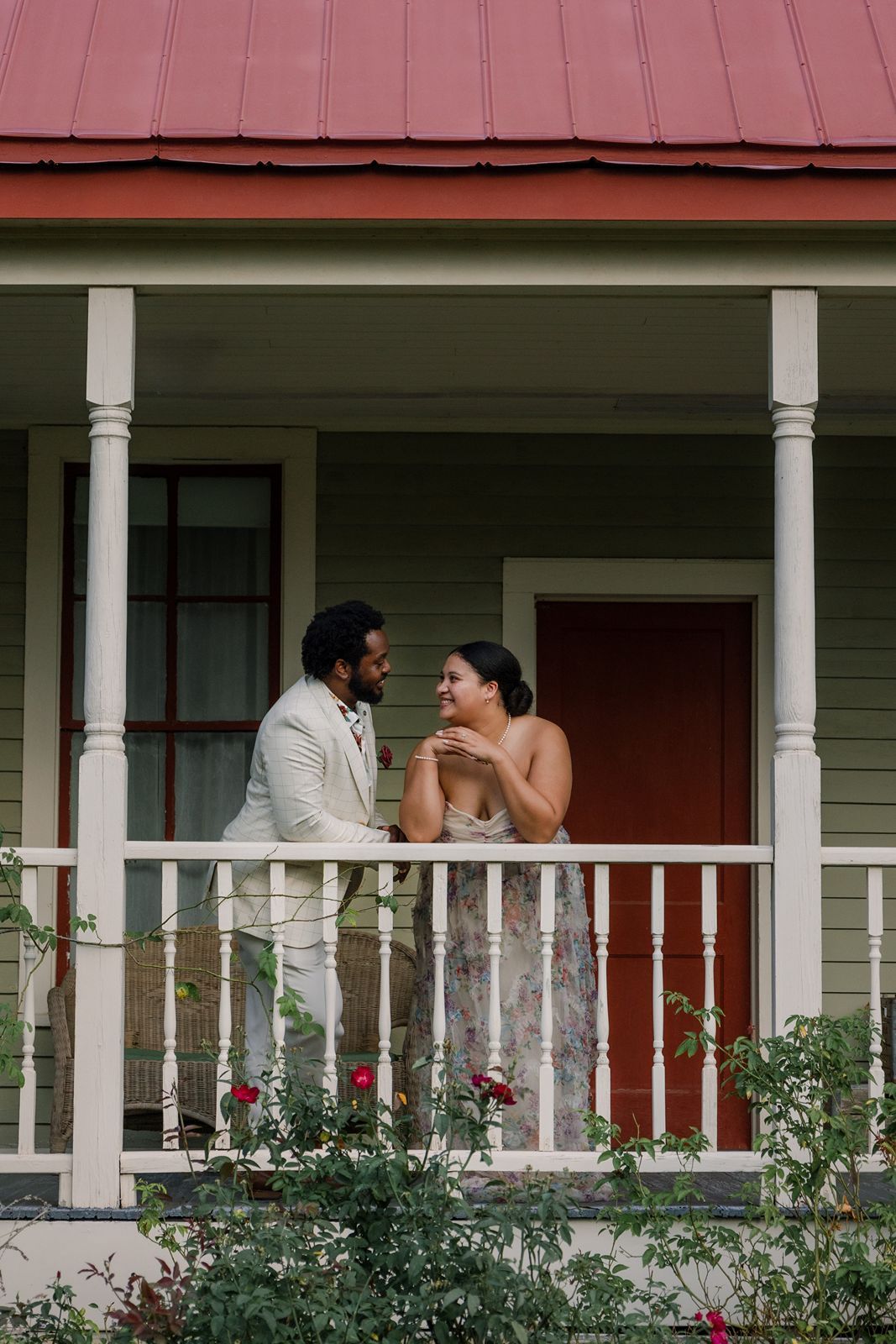 A couple shares a romantic moment on a front porch with white columns and green bushes below.