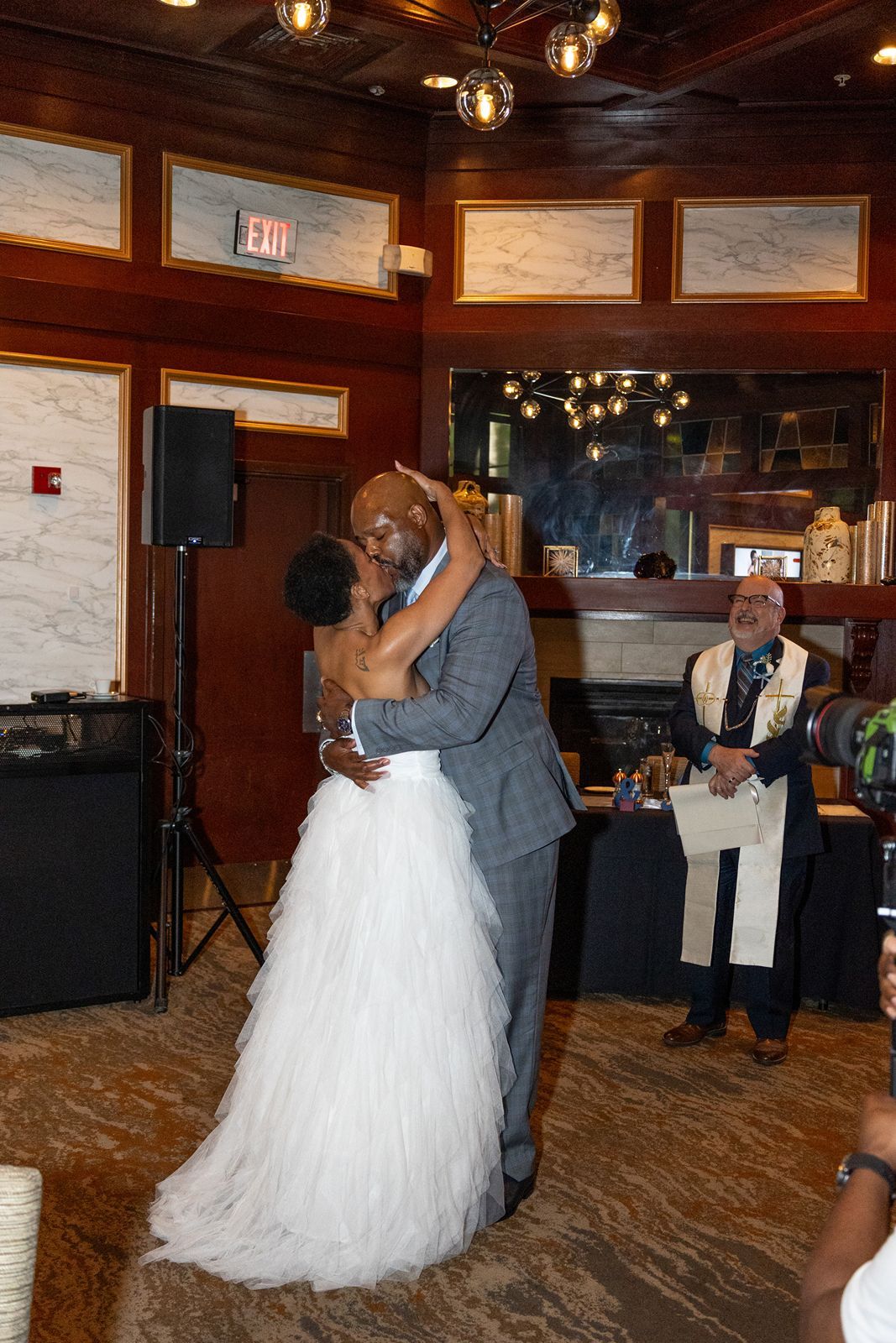 Newlyweds share first kiss as husband and wife while officiant watches during indoor wedding ceremony.