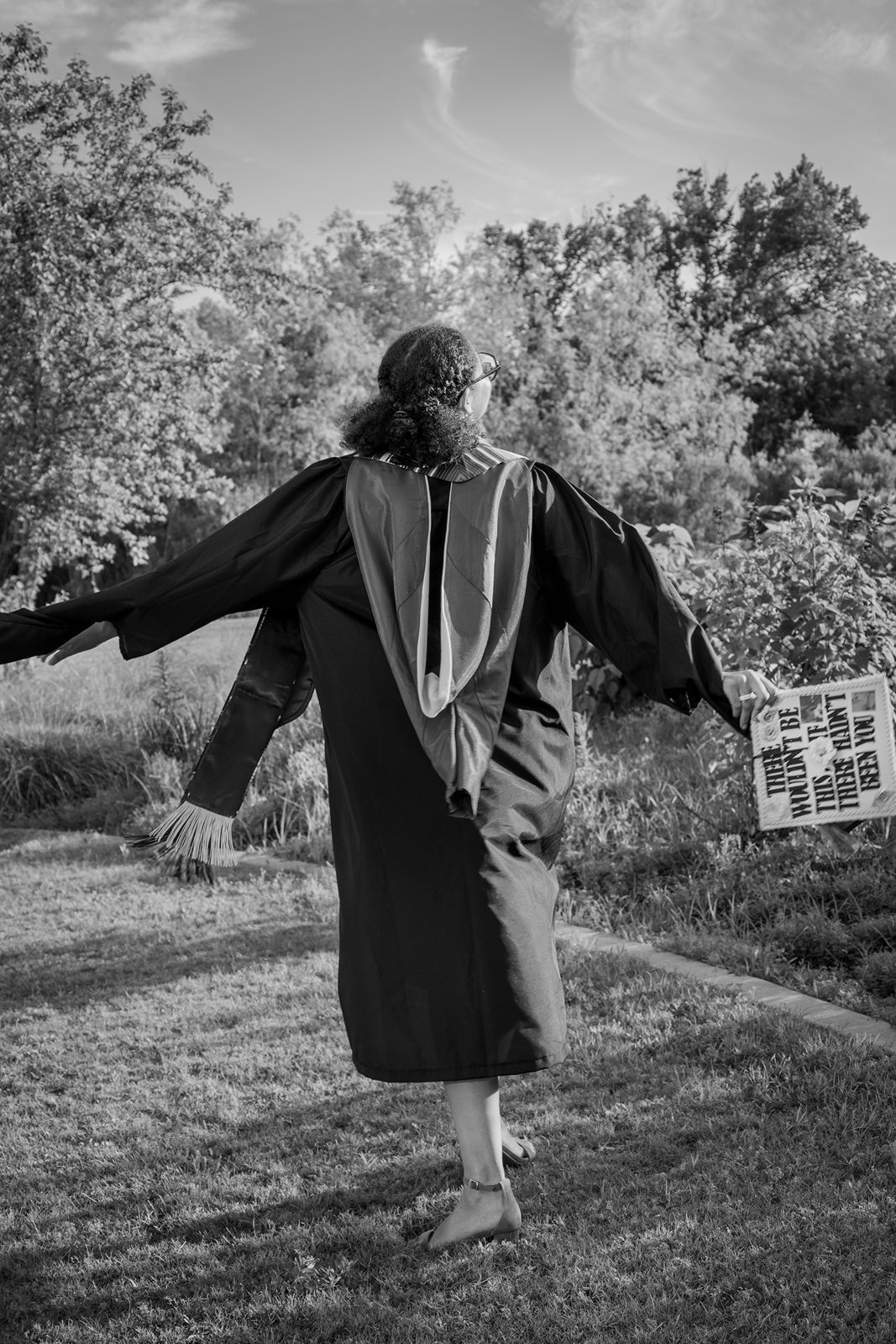 Graduate in black gown walks outdoors holding diploma, black and white photo with trees in background.