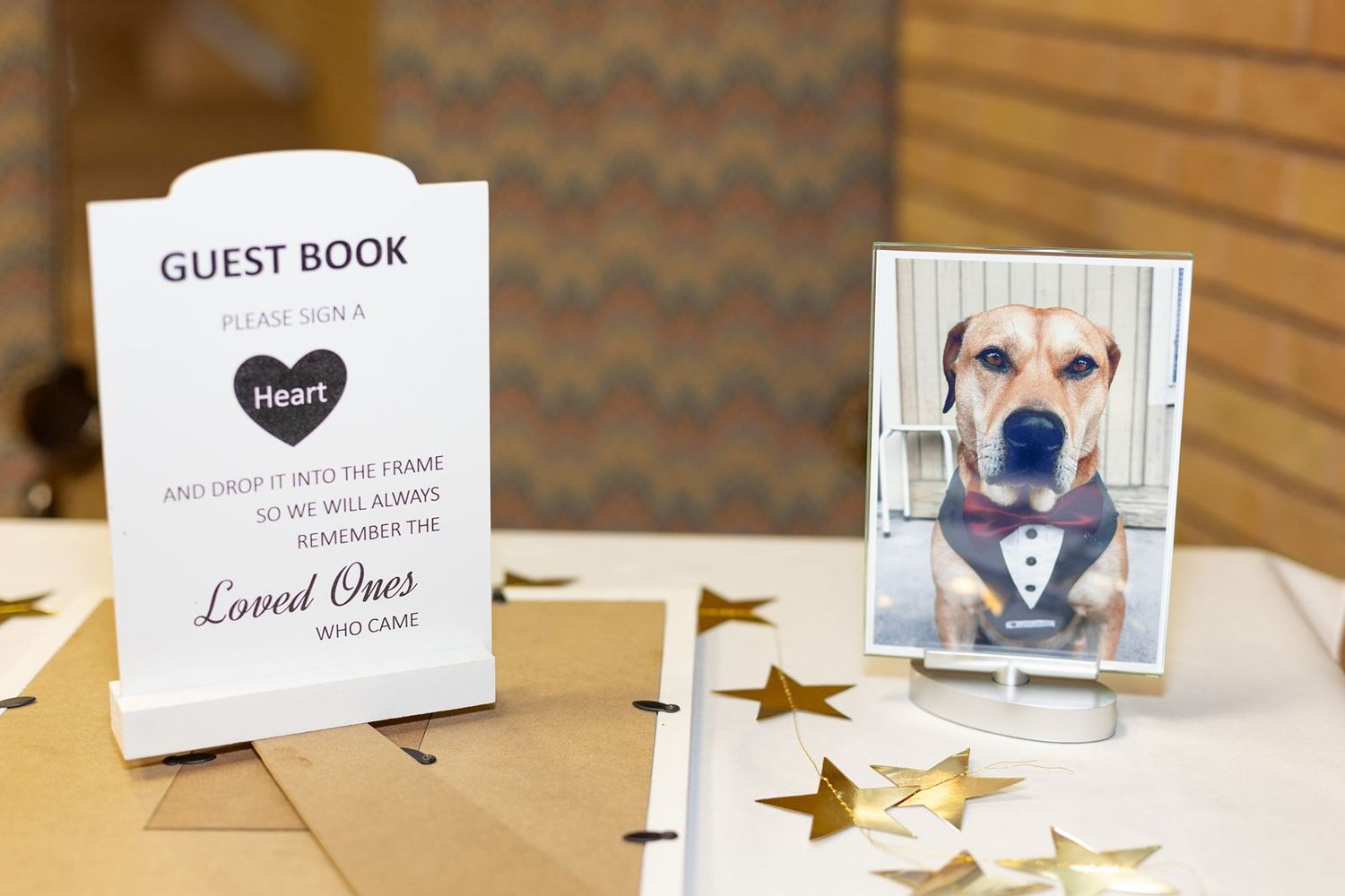 Wedding guest book sign beside a framed photo of a dog in a tuxedo on a table with gold star confetti.