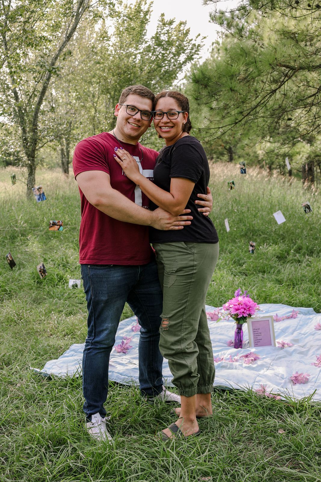 Happy couple embracing on a picnic blanket outdoors, surrounded by greenery and pink flowers.