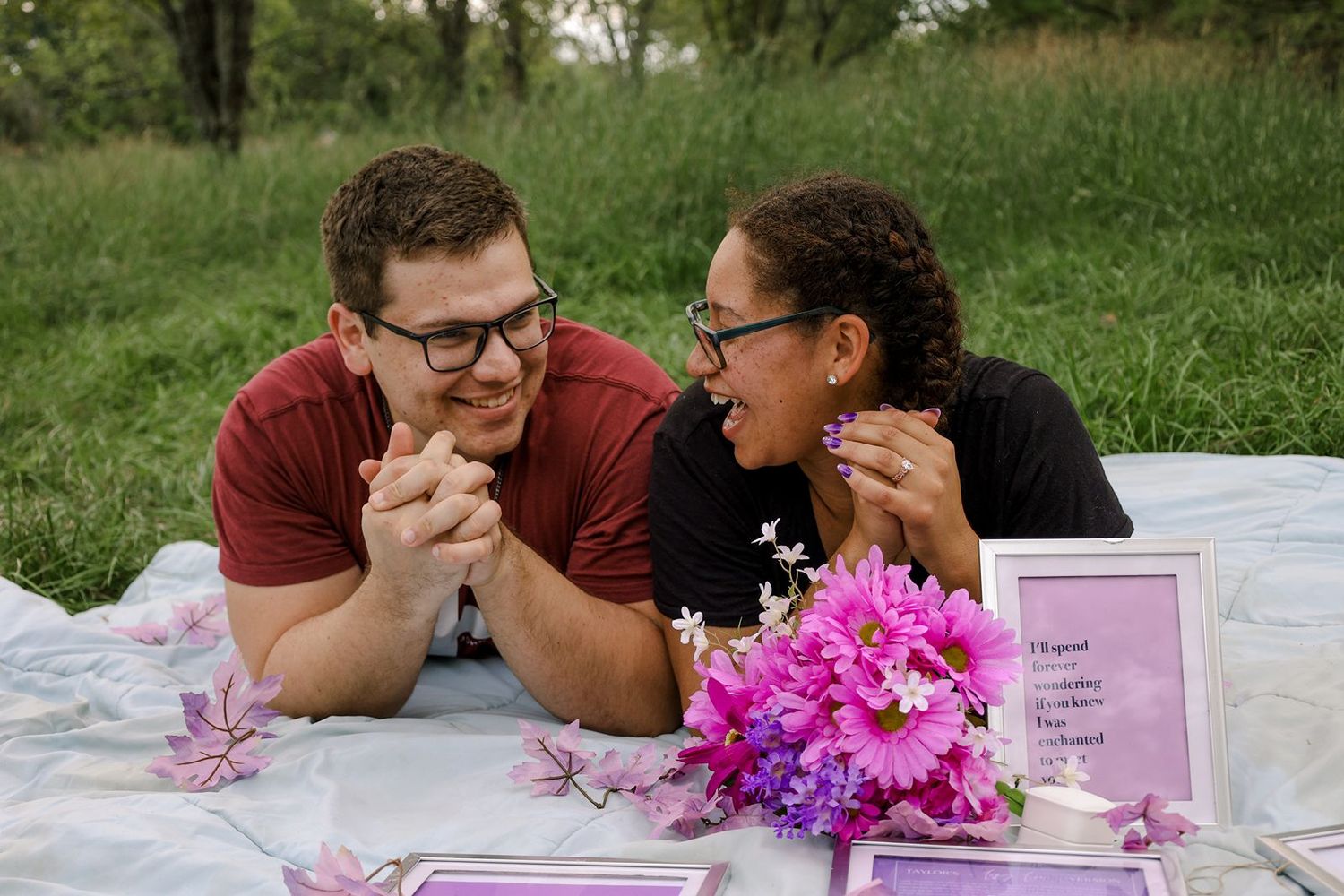 A newly engaged couple laughs joyfully together on a white blanket with pink flowers and framed notes.
