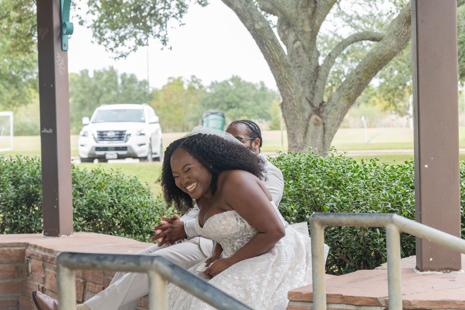 Newlyweds share playful moments together on a brick ledge with trees in the background.