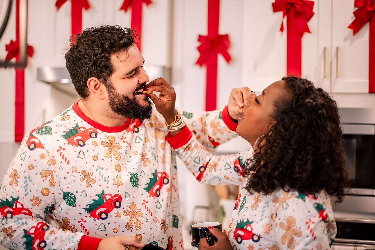 Two people in matching holiday pajamas share a joyful moment in a kitchen decorated with red holiday decor.