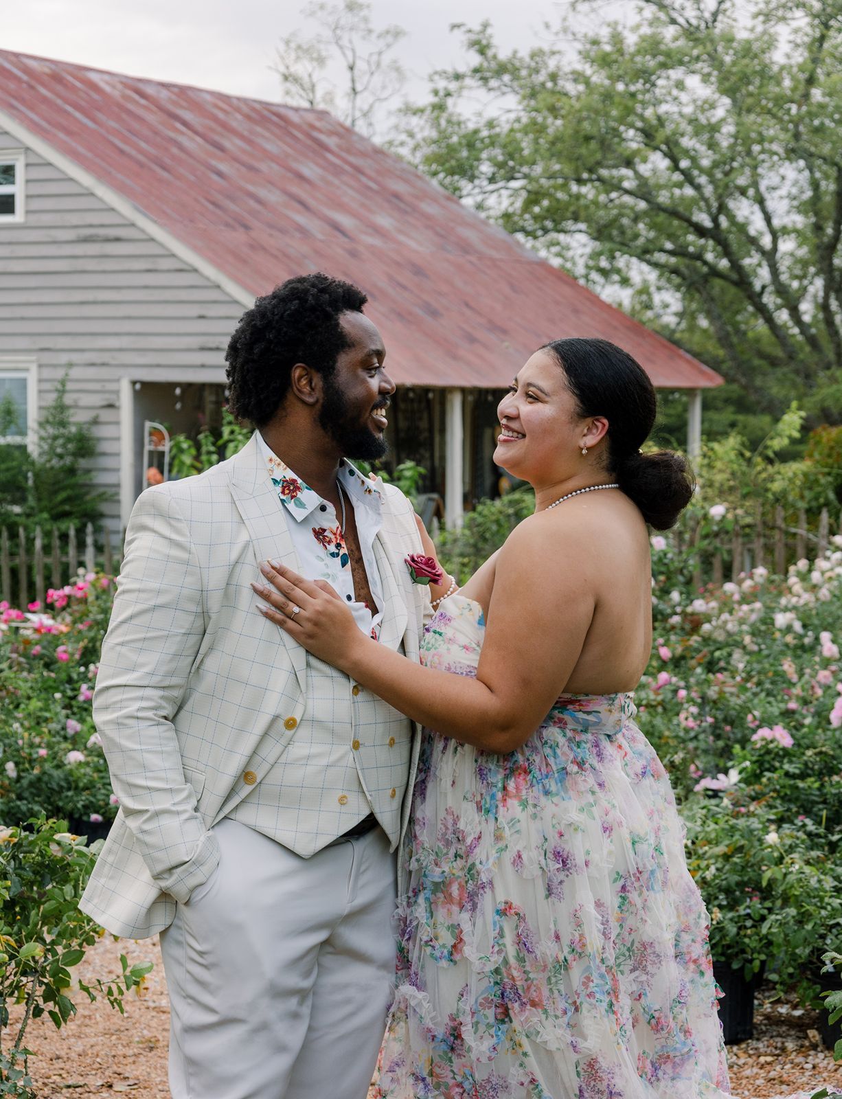 A couple shares joyful moments together in a garden setting with blooming roses and rustic buildings in the background.