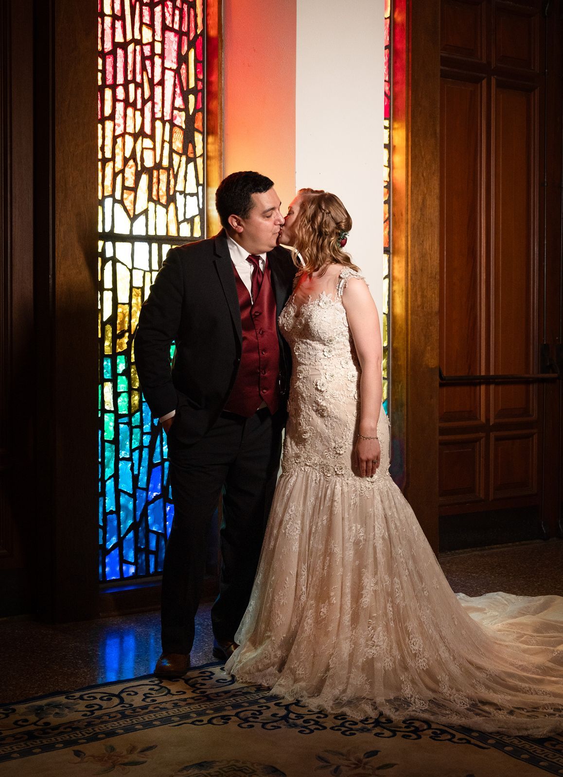A couple shares an intimate moment in front of a stained glass window casting colorful light in a church setting.