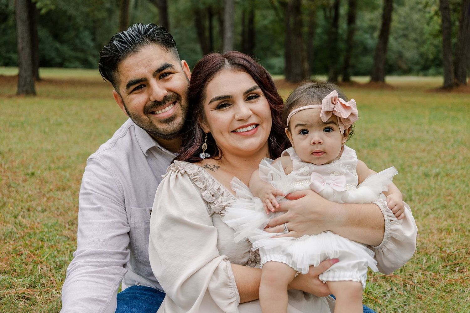 A family poses together outdoors in coordinated white outfits against a green wooded background.