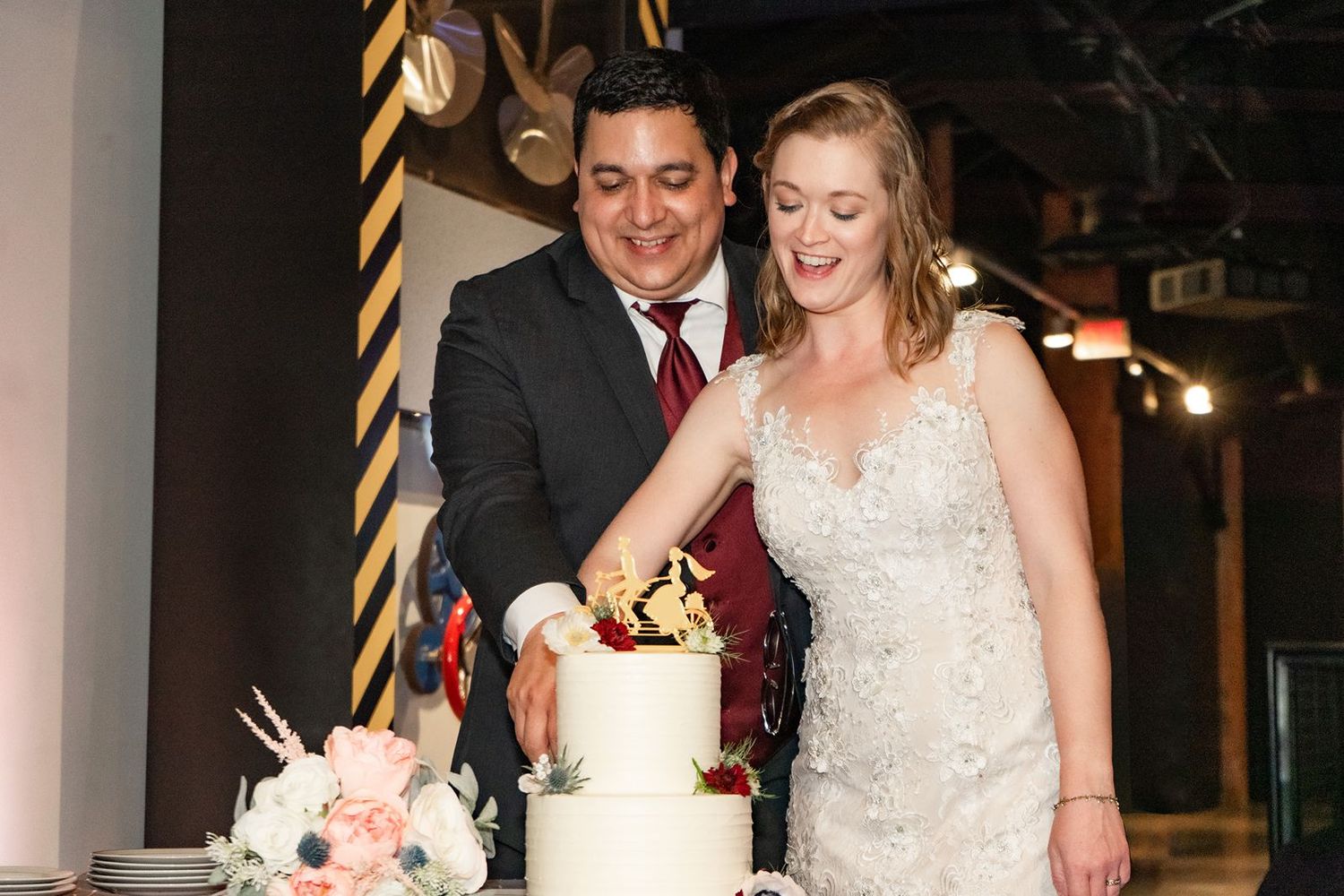Bride and groom smile while cutting their white two-tier wedding cake at their reception.