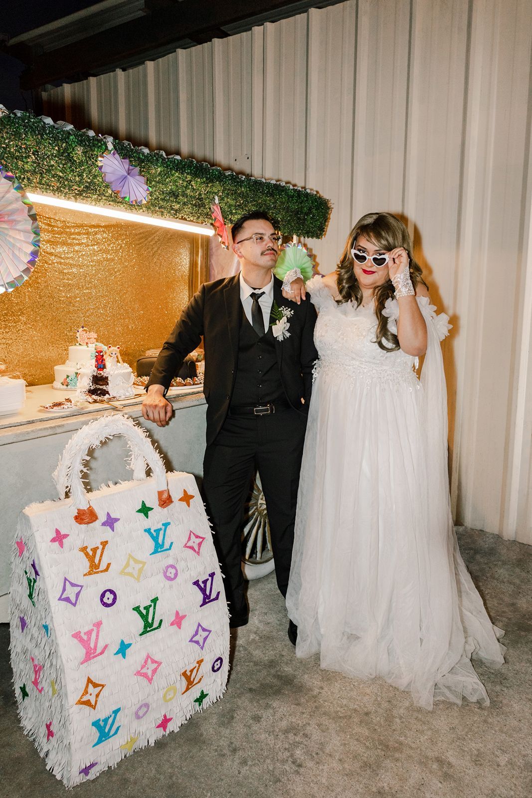 Bride in white gown with fun sunglasses and groom in black suit pose with a Louis Vuitton piñata at wedding reception.