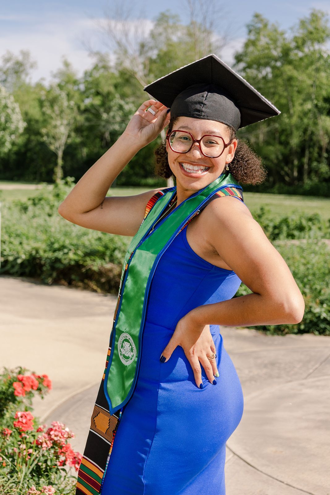 A person wearing a blue dress and graduation cap poses outdoors near pink flowers on a sunny day.
