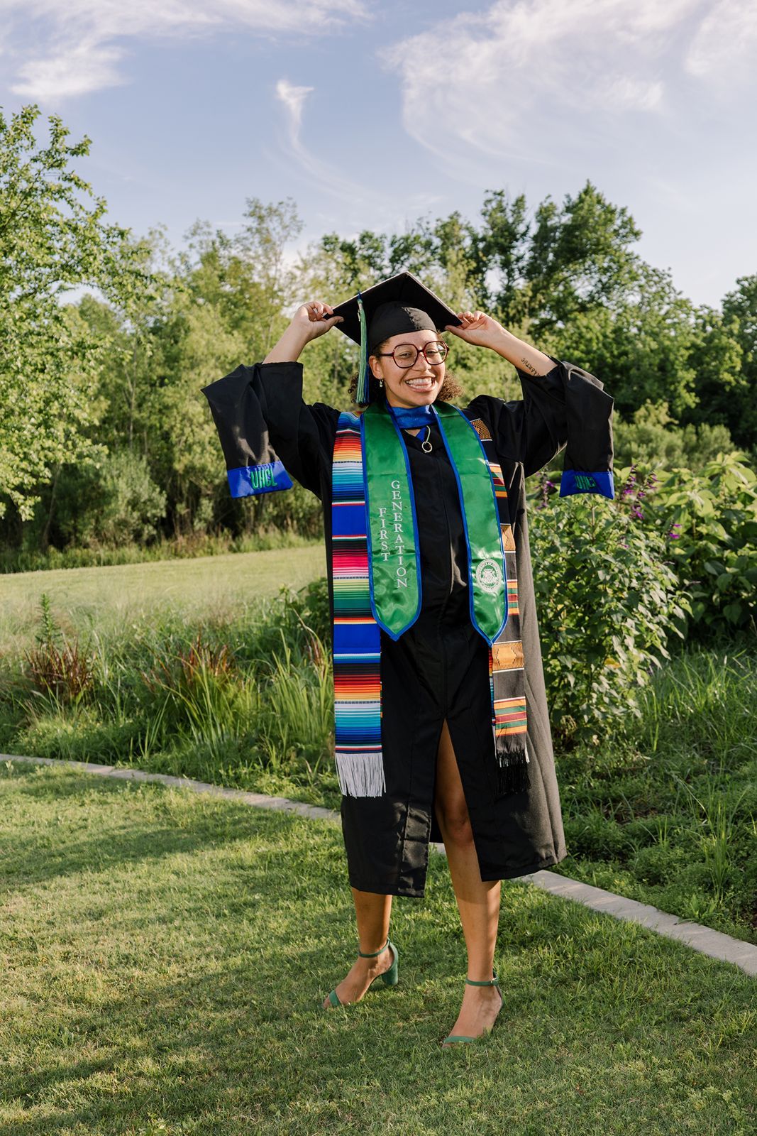 Graduate in cap and gown poses outdoors with colorful graduation stoles against green natural background.