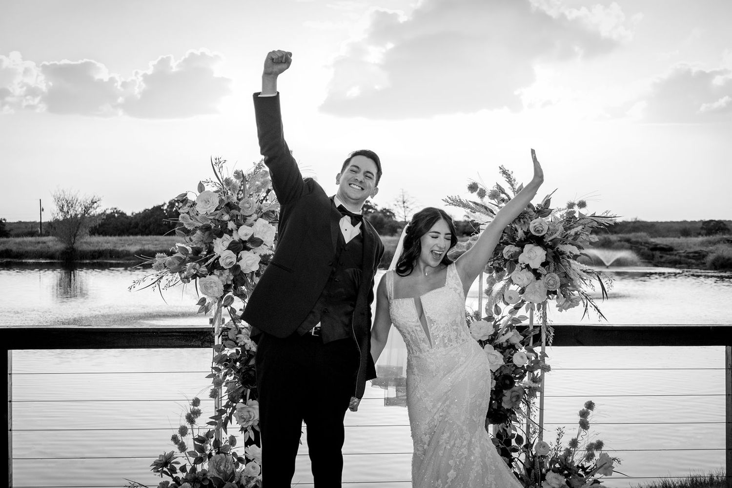 Bride and groom celebrating joyfully in black and white photo with floral arch at waterfront venue.