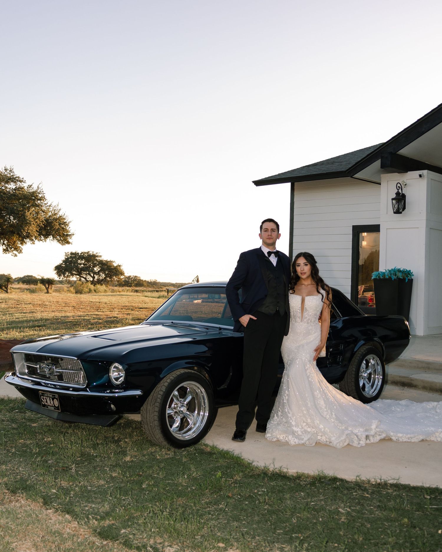 Bride and groom posing beside a classic black Ford Mustang at a rustic Texas wedding venue at golden hour.