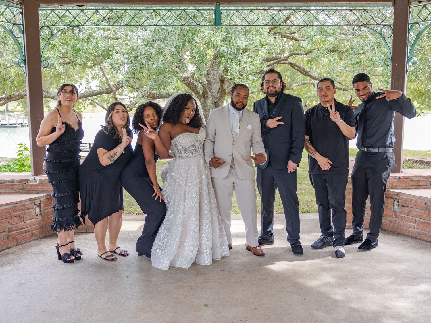 Wedding party members pose together in coordinated formal attire under a covered brick pavilion.