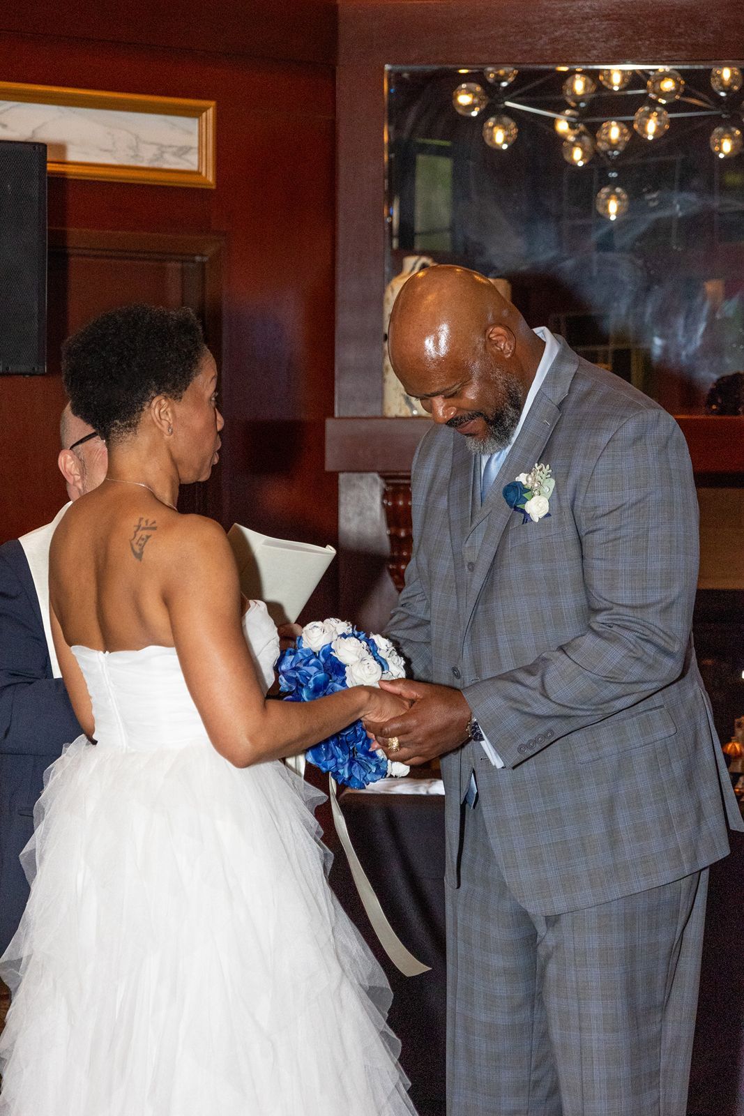 Groom in gray plaid suit places wedding ring on brides finger during indoor ceremony.