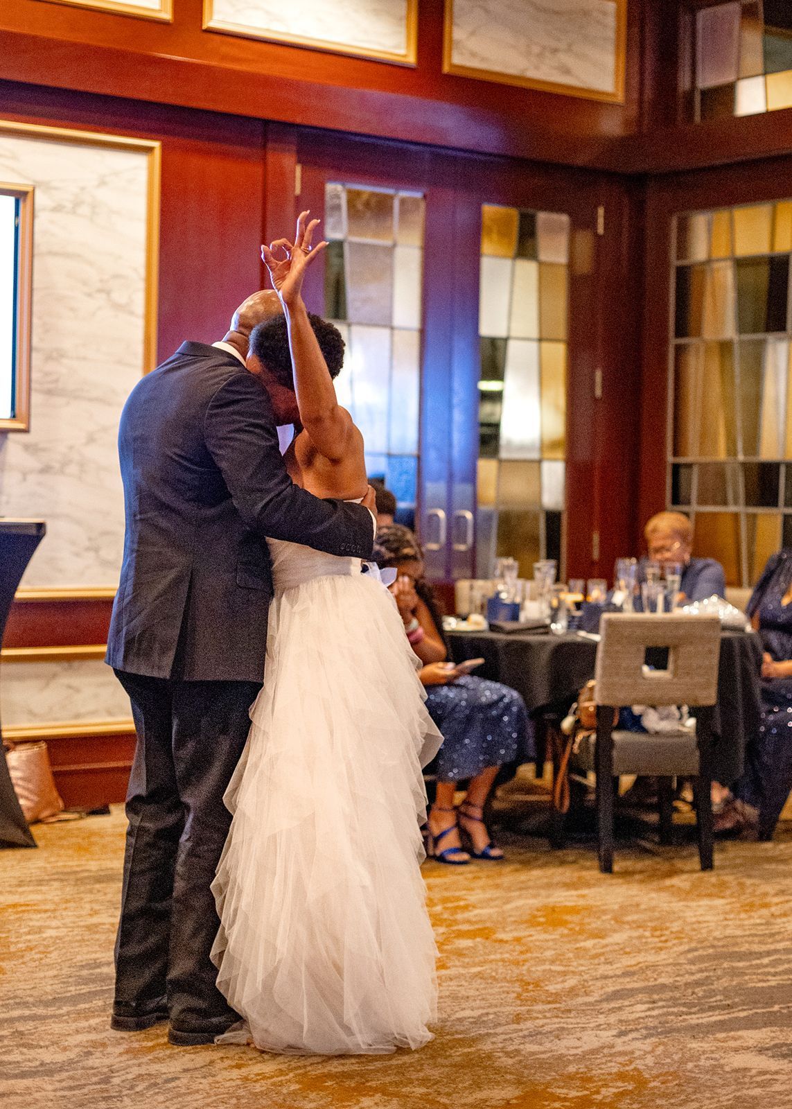 Bride and groom share their first dance at elegant wedding reception venue with guests seated nearby.