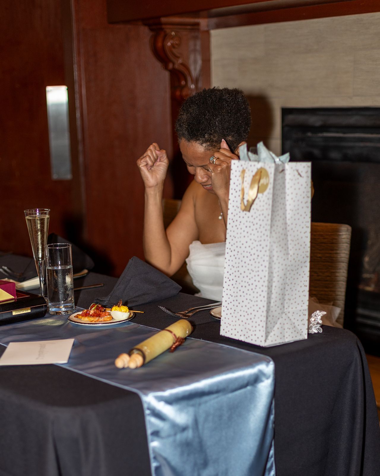 Bride in white strapless gown sits at decorated reception table with gift bag and food.