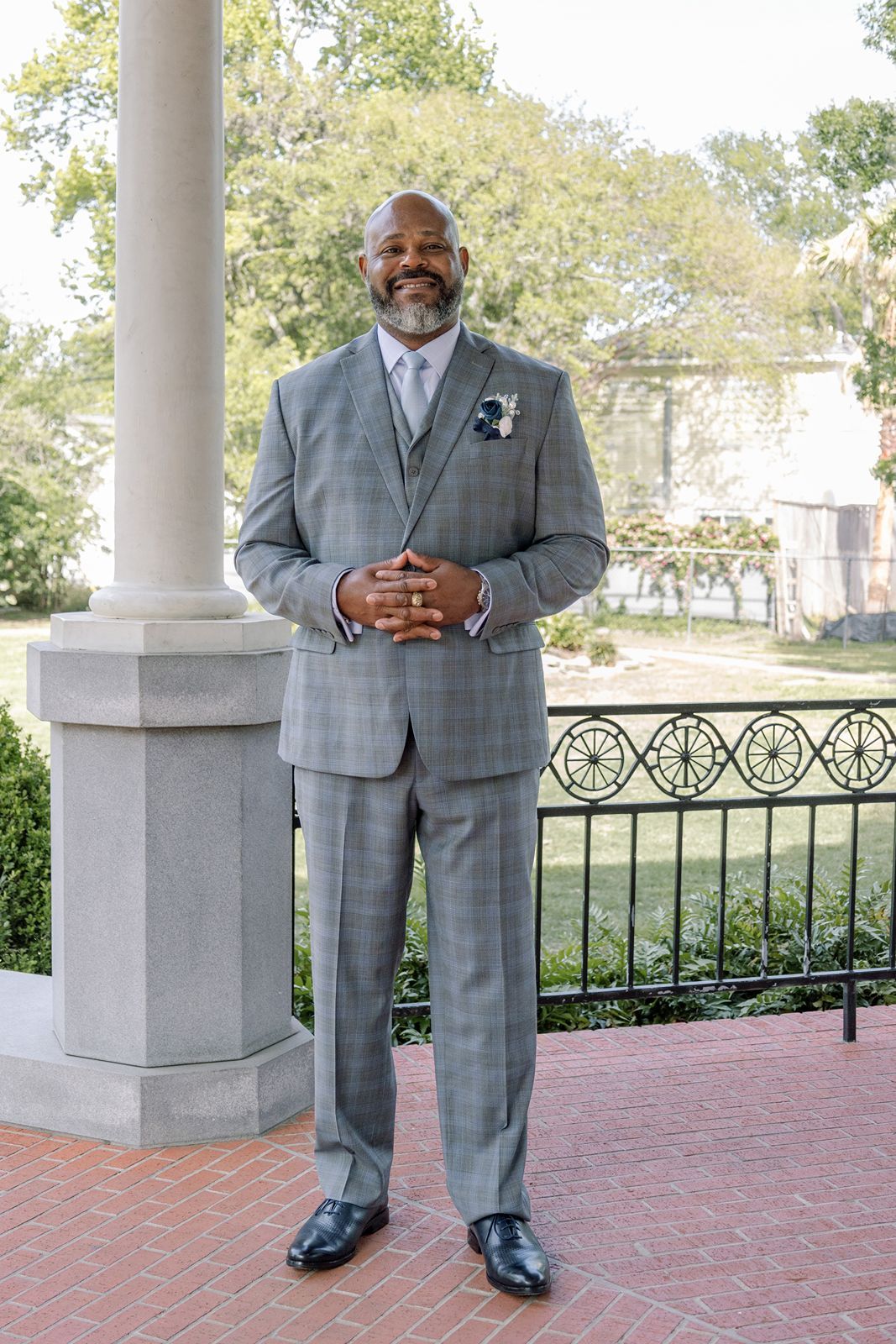 Dapper groom in gray plaid three-piece suit with boutonniere standing on brick porch outdoors.