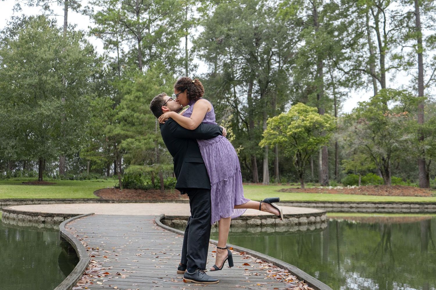 Couple embracing and lifting near a reflective pond on a curved wooden walkway in a lush park.