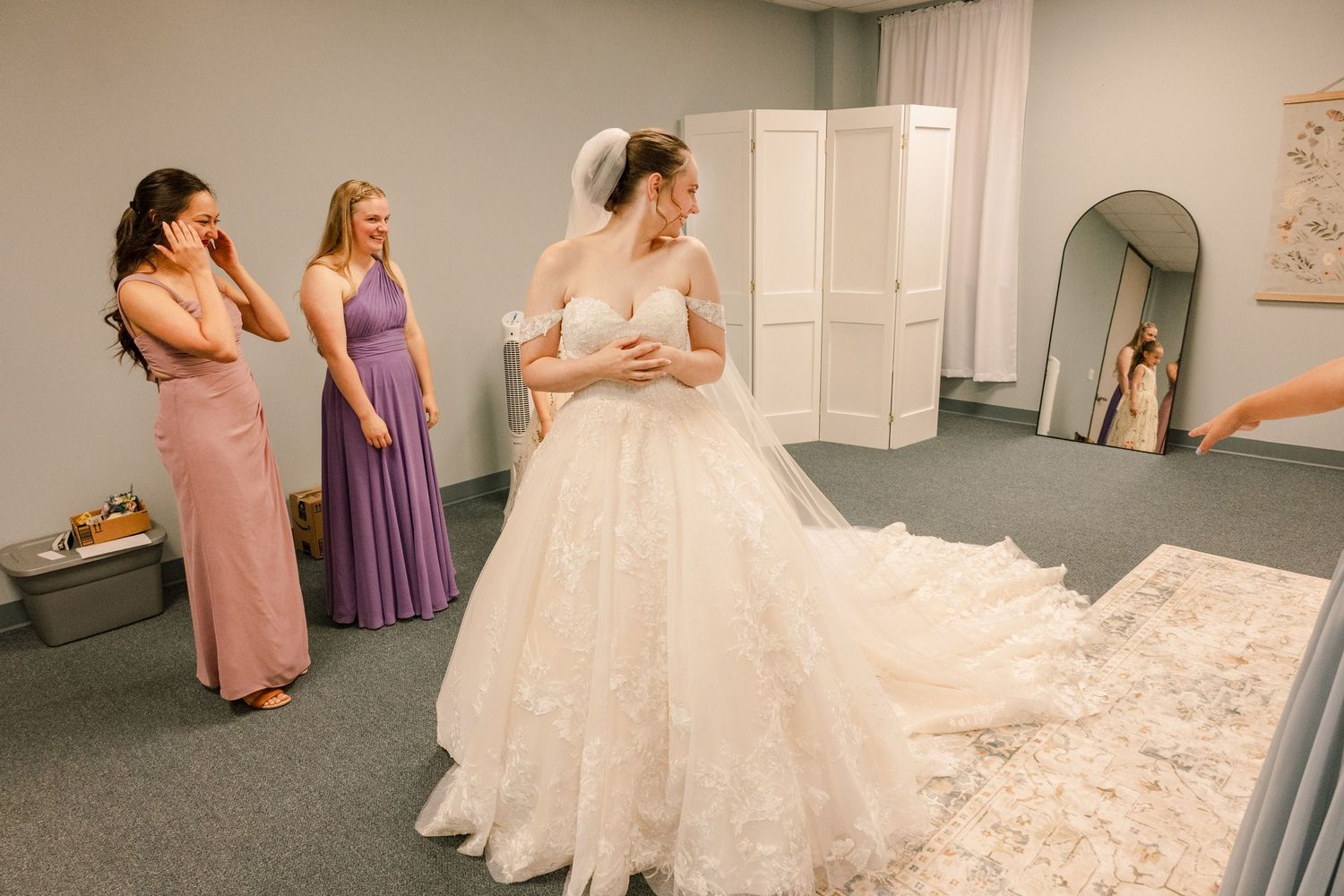 Women in pastel dresses help arrange a long wedding dress train on gray carpet.