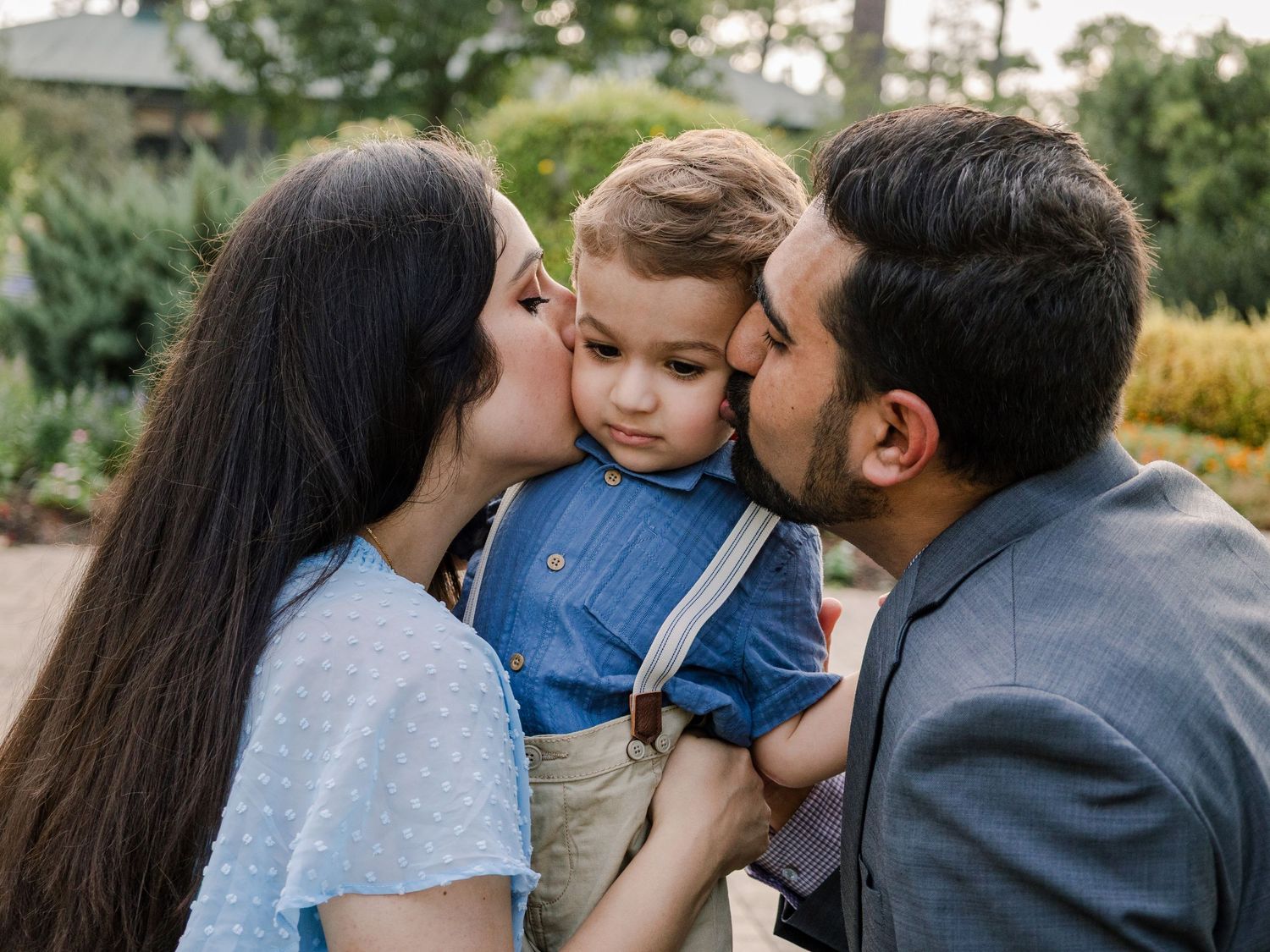 A couple in casual and formal attire sharing a tender moment with a young child in denim overalls outdoors.