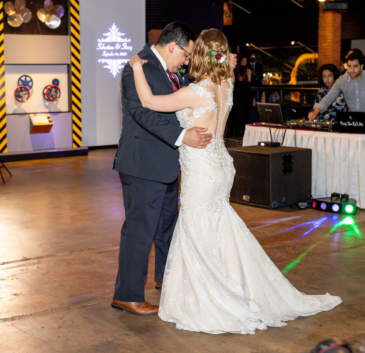 Bride and groom share their first dance at wedding reception with DJ booth visible in background.