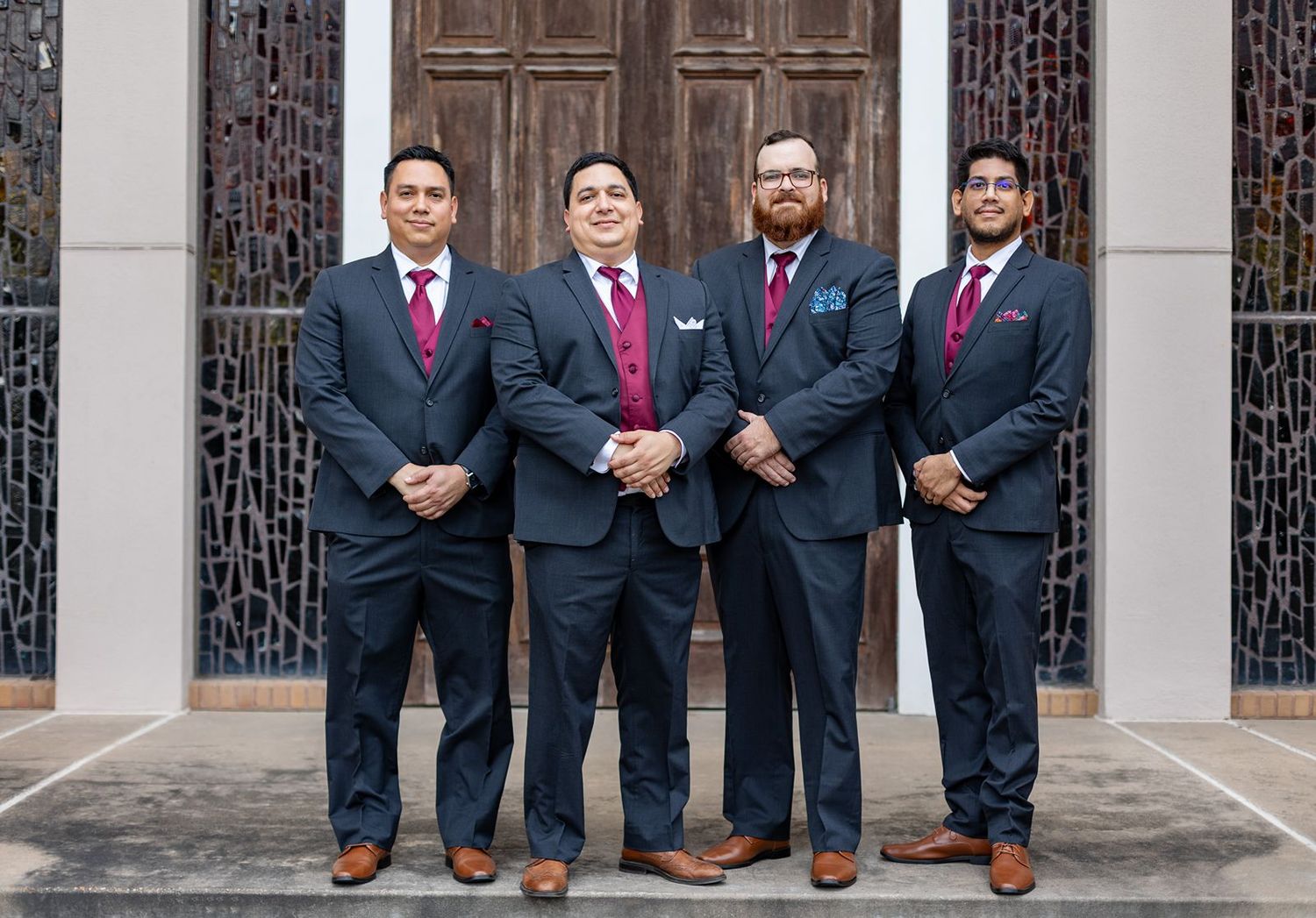 Four men in matching dark suits with burgundy ties pose formally outside a decorative church entrance.