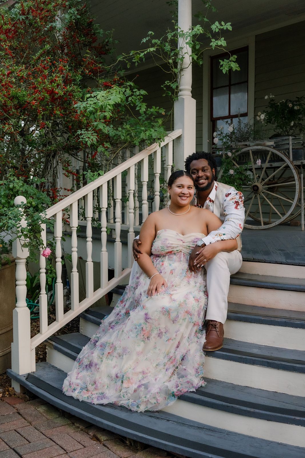 A couple sits together on gray porch steps with white railings and greenery in the background.