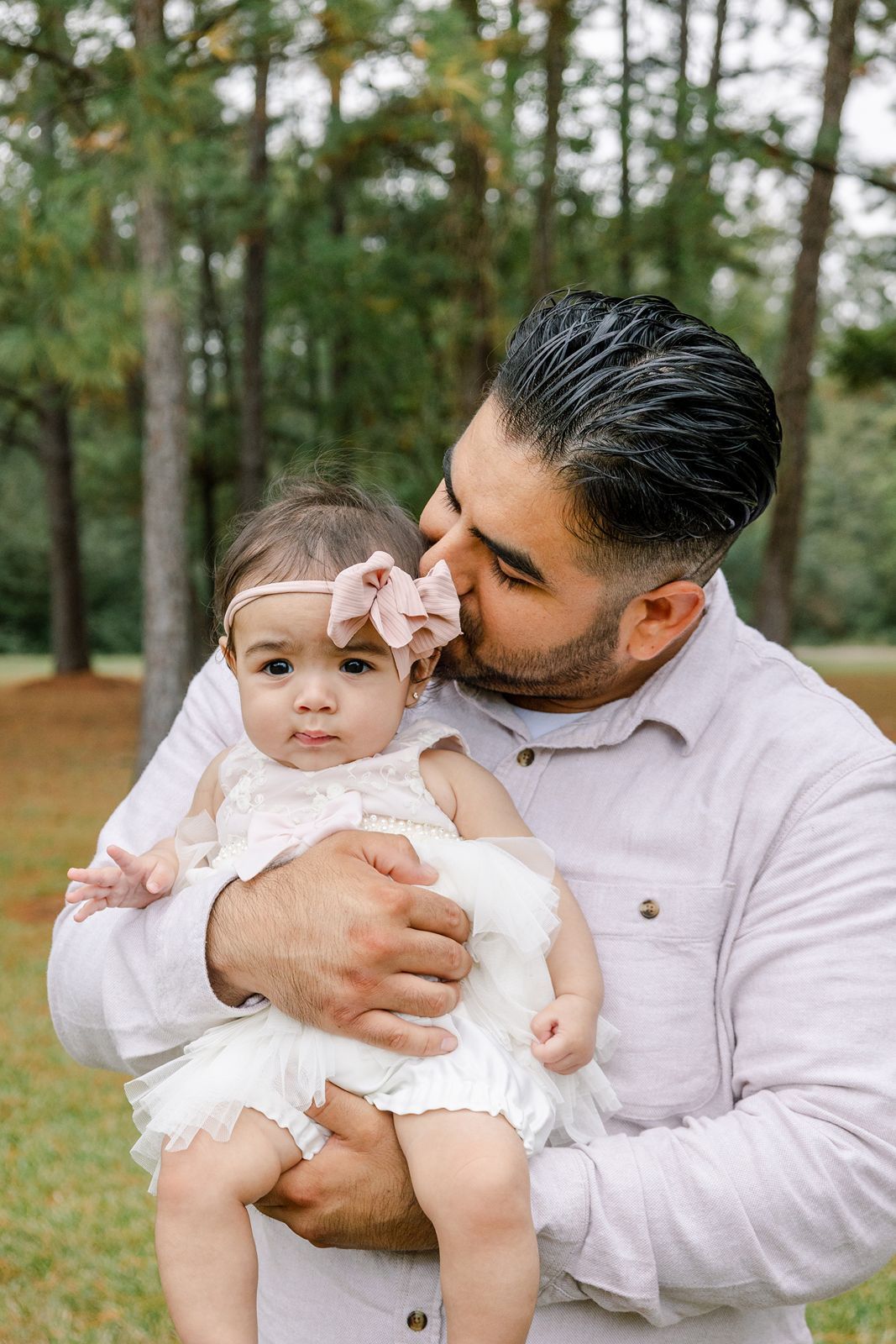 A tender moment between parent and baby in white outfits against a wooded outdoor setting.