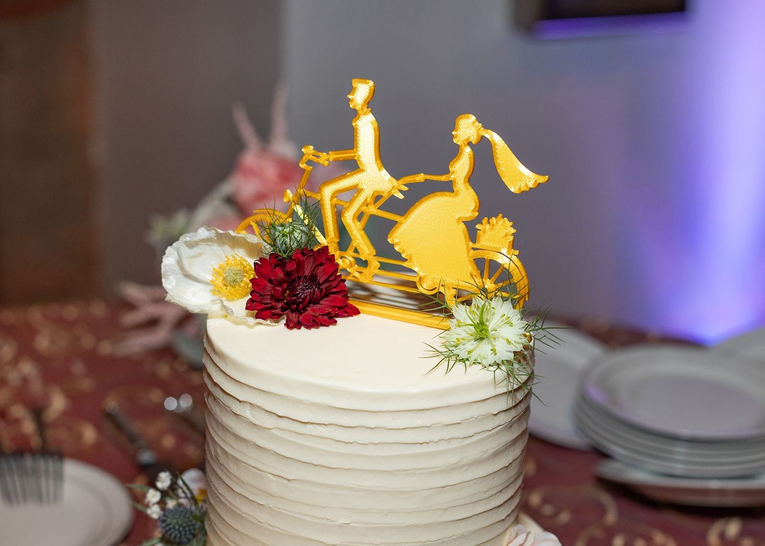 White wedding cake with gold bicycle topper and red flowers on a decorated table.